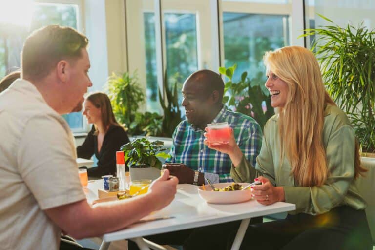 woman seated on a chair during lunch with two more person. She is holding a bevarage, and they are eating a meal on a office cantine.